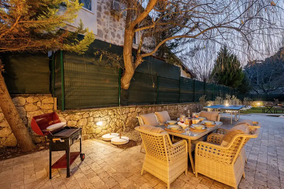 An outdoor dining table and grill under a tree at dusk, lit by string lights