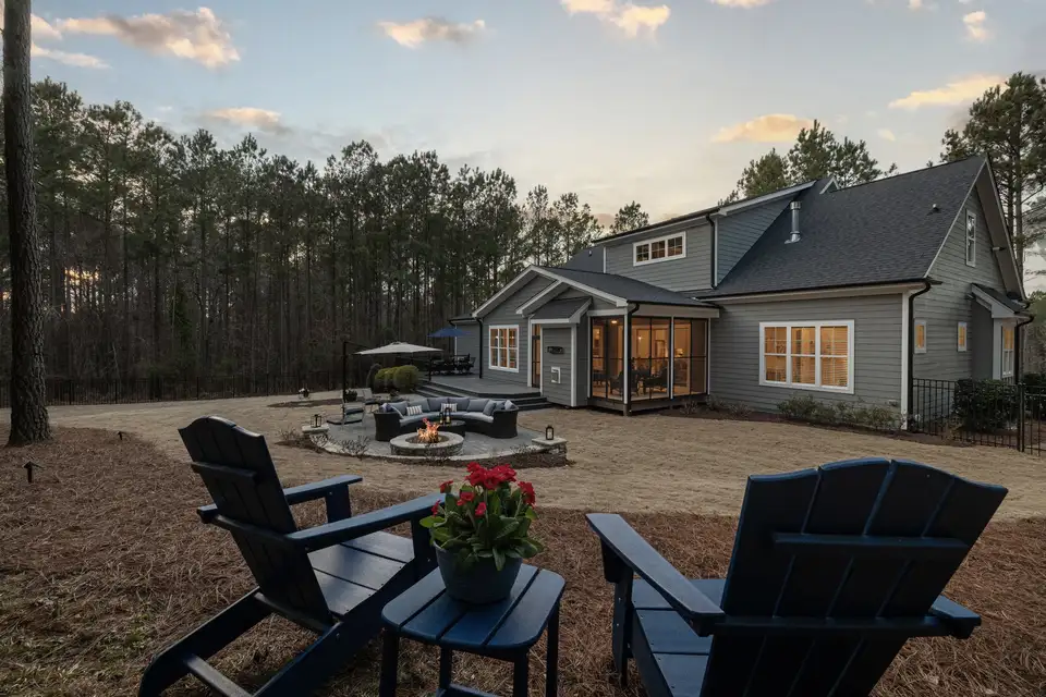A backyard fire pit with Adirondack chairs and a lit house at dusk